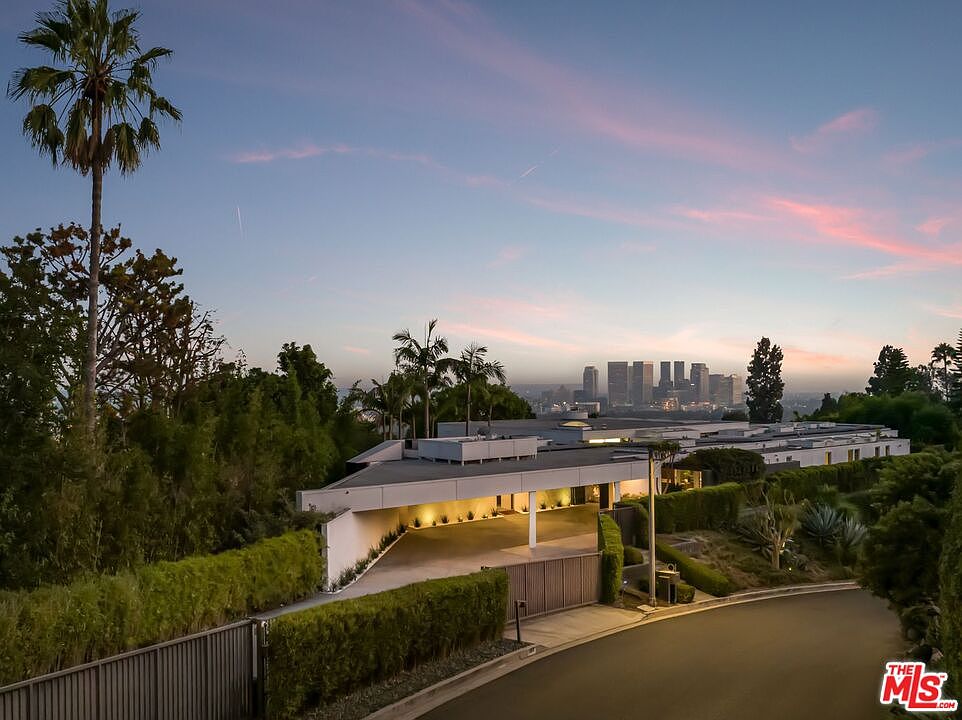 This image showcases the front exterior of a modern, sprawling residence at dusk. The property features a long, low-slung design with a flat roof, a gated entrance, and lush landscaping that provides privacy. In the background, the city skyline adds a touch of urban sophistication to the serene setting.