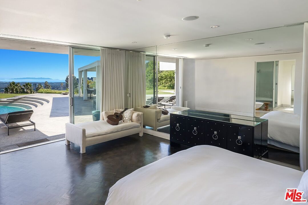 This is a bright and modern primary bedroom with floor-to-ceiling windows offering views of the pool and landscape. The room features a large bed with white linens, a sleek black dresser with mirrored accents, and a white chaise lounge. The dark wood floors add a touch of warmth to the otherwise minimalist design.
