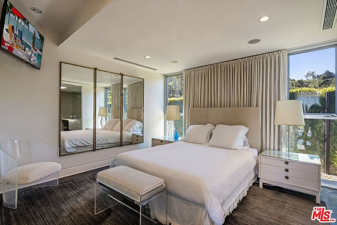 This primary bedroom features a king-sized bed with white linens, flanked by matching nightstands and lamps. A unique three-panel mirror reflects the bed and part of the room, adding depth. The room is well-lit with natural light from a window with curtains, and a TV is mounted on the wall.