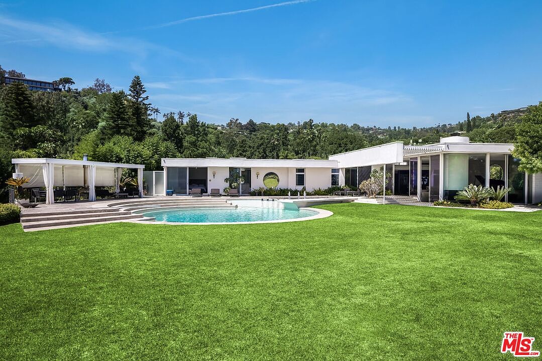This image showcases the rear exterior of a modern home, emphasizing the expansive green lawn and a circular swimming pool. The house features a sleek, white facade with large windows and a unique circular window detail. A covered patio area provides an outdoor living space, and lush greenery surrounds the property, creating a private and luxurious atmosphere.