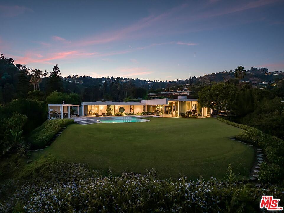 This aerial view showcases a stunning modern home with a sprawling green lawn, a circular pool, and lush landscaping. The house features clean lines, large windows, and a flat roof, blending seamlessly with the surrounding hillside. The property exudes luxury and privacy, making it an ideal retreat.