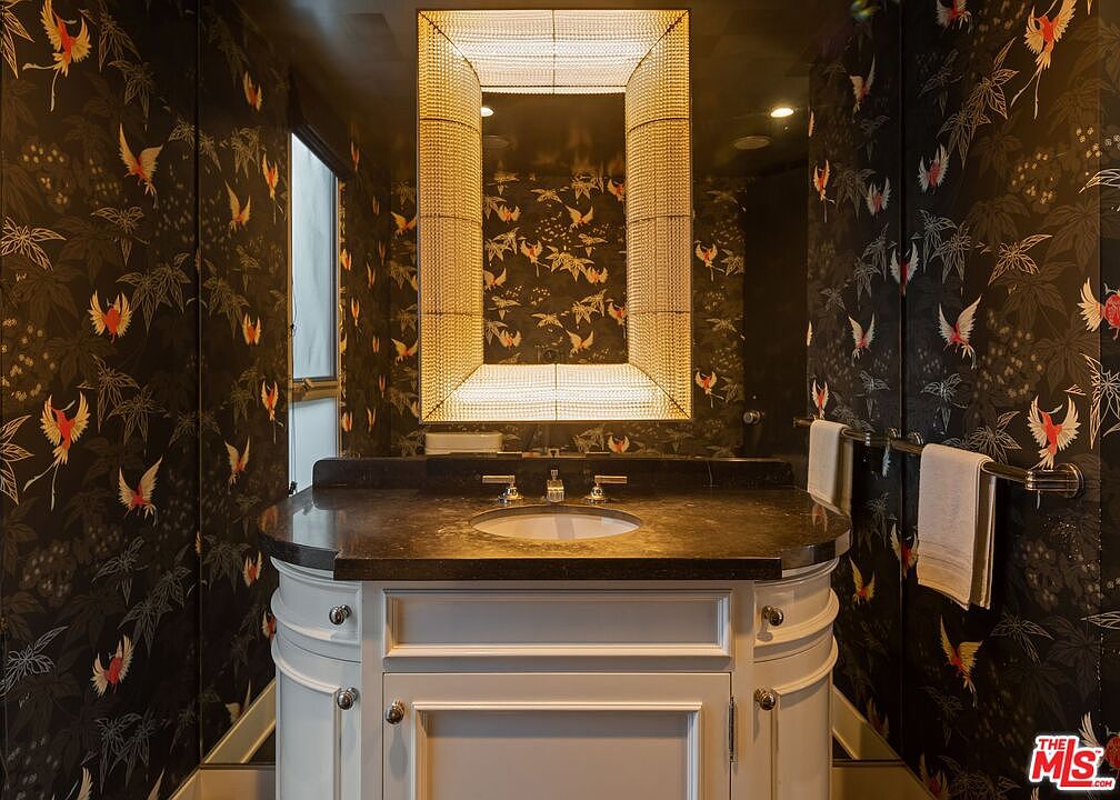 This is a stylish guest bathroom featuring dark, patterned wallpaper with bird and leaf motifs. A white vanity with a dark countertop and a unique, illuminated mirror create a focal point. The overall impression is luxurious and sophisticated, with a touch of whimsy.