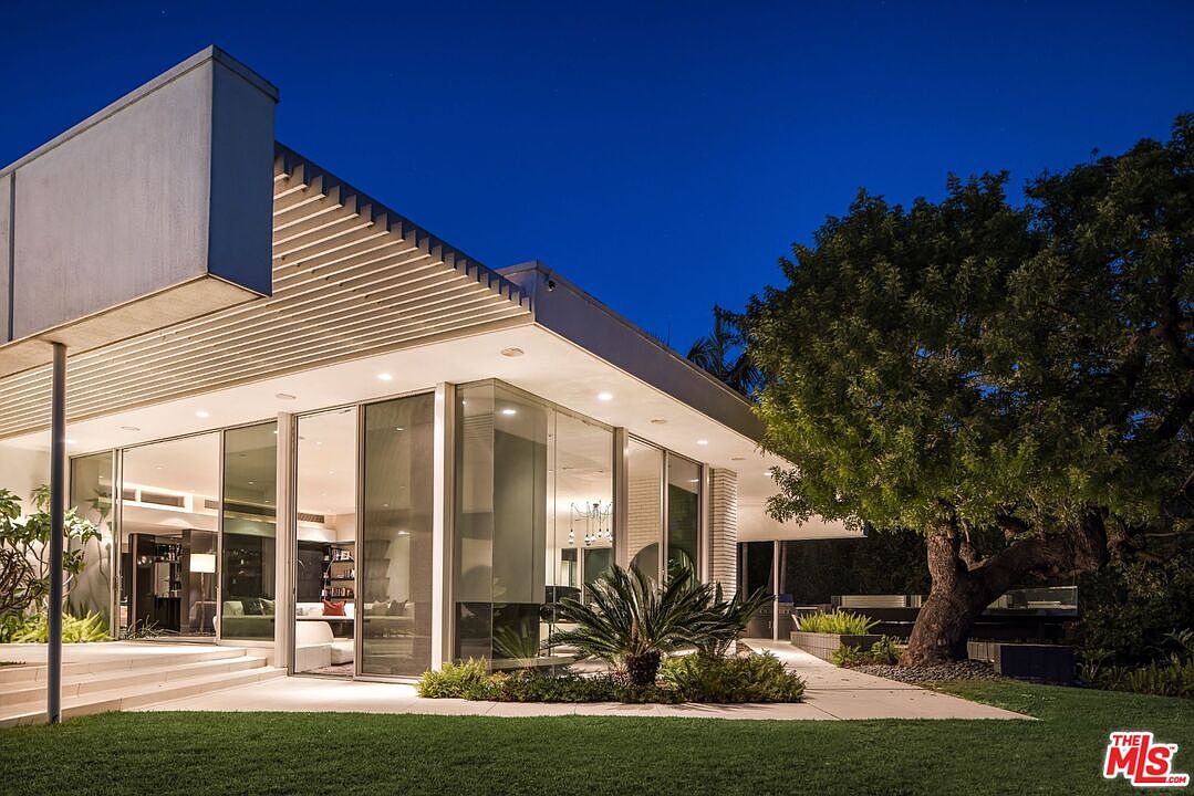 This is a front exterior view of a modern home at dusk. The house features large glass windows and doors, allowing a glimpse into the interior living spaces. A well-manicured lawn and mature trees add to the property's curb appeal, while the architectural design emphasizes clean lines and a minimalist aesthetic.