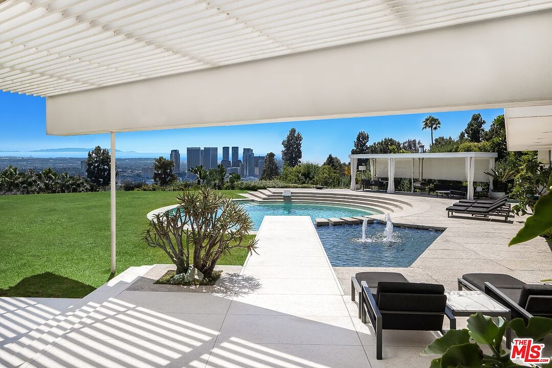 This image showcases a luxurious outdoor pool and spa area with a stunning city view. The pool features a unique design with a connecting water feature, surrounded by a spacious patio with modern lounge furniture. The well-manicured lawn and architectural details of the covered patio enhance the property's appeal, creating an inviting and upscale outdoor living space.