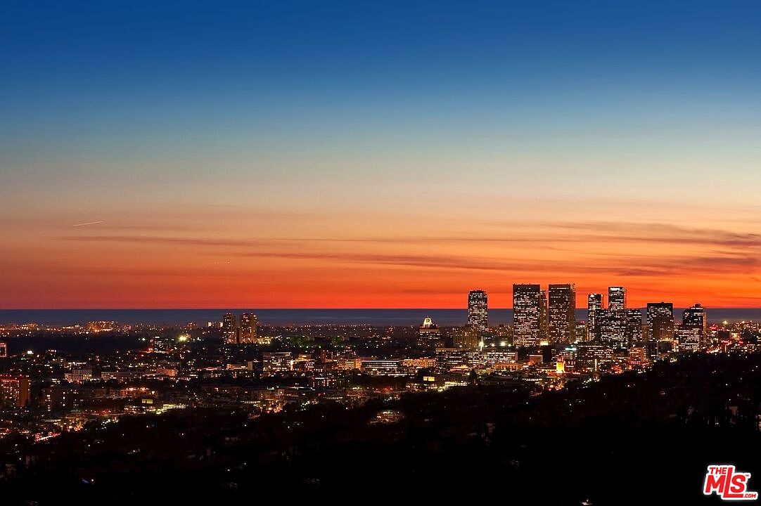 This image showcases a stunning aerial view of a city skyline at dusk, with the sky transitioning from deep blue to vibrant orange. The city lights twinkle against the darkening landscape, highlighting the architectural details of the buildings. The overall impression is one of urban sophistication and scenic beauty, making it an attractive feature for a real estate listing.