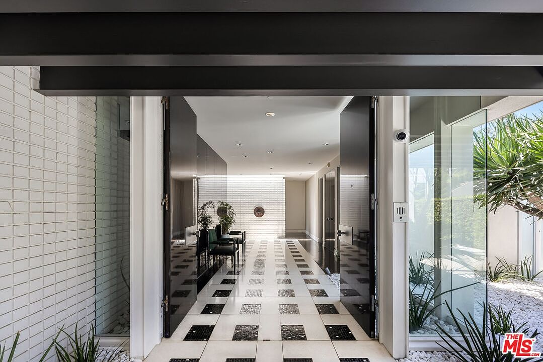This image showcases a stylish entryway with a striking black and white tiled floor leading into a modern home. The entrance features large, dark-framed doors and a glass wall that allows natural light to flood the space, highlighting the minimalist design and clean lines. The exterior wall is clad in white tiles, adding texture and visual interest.