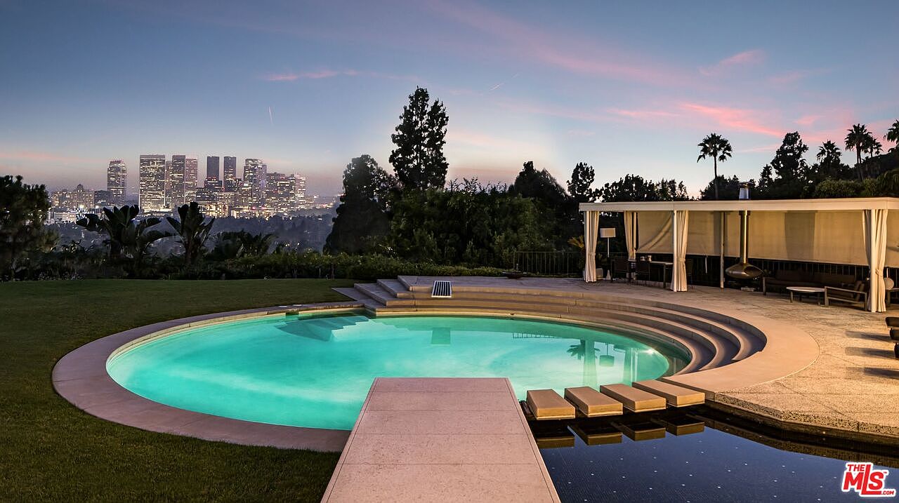 This image showcases a luxurious backyard pool area at dusk, featuring a circular pool with a modern design and a connecting walkway. The pool is surrounded by lush greenery and a covered patio area with seating. In the background, the city skyline adds a touch of urban sophistication to the serene outdoor setting.