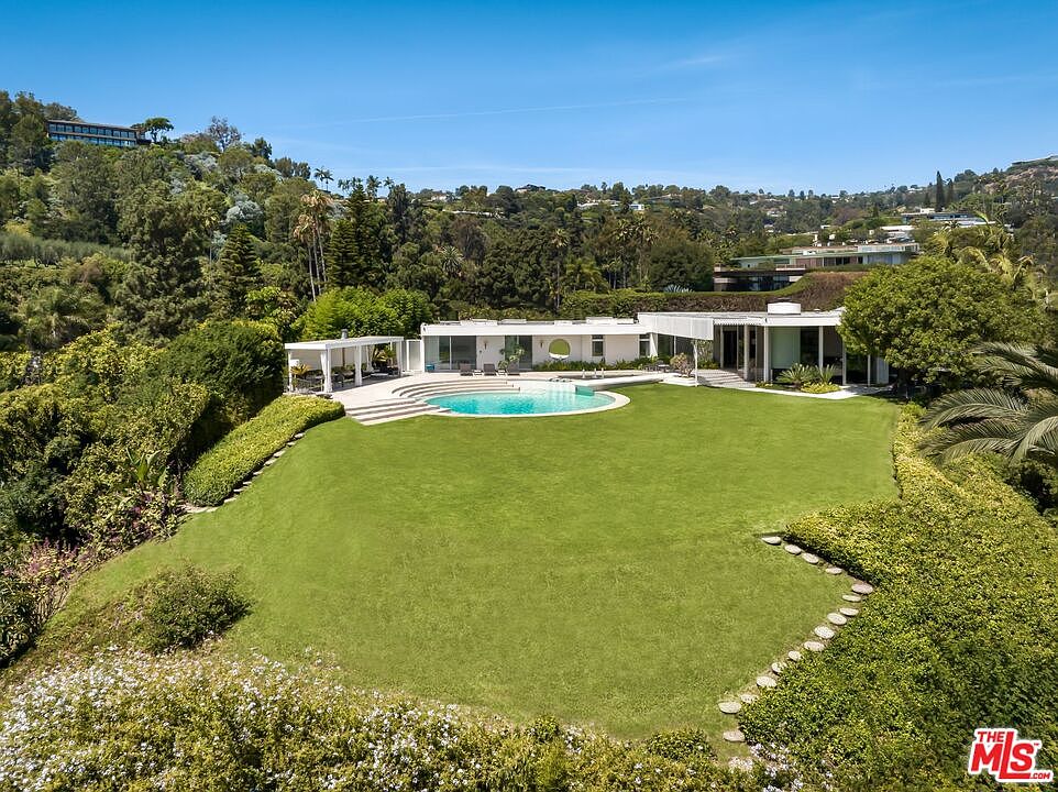 This aerial shot showcases a modern, single-story home with a flat roof, surrounded by lush greenery and mature trees. A circular pool is a focal point, complemented by a large, well-manicured lawn. The property exudes a sense of privacy and luxury, blending seamlessly with the natural landscape.