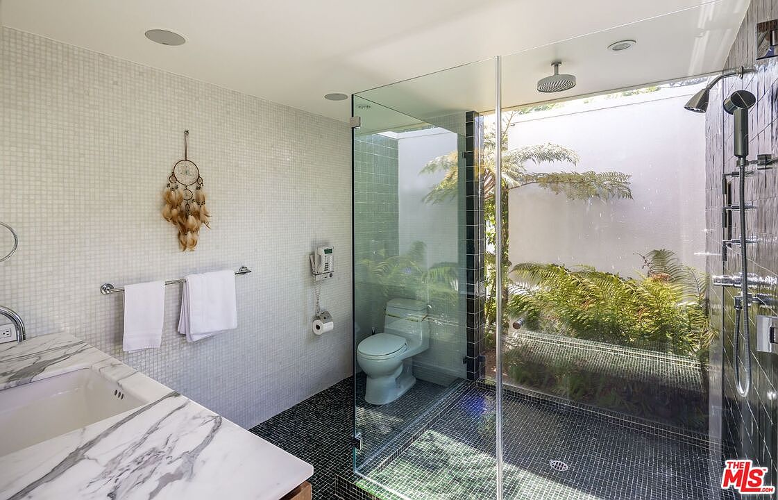 This is a bright and modern primary bathroom featuring a glass-enclosed shower with a view of an outdoor garden. The bathroom has a double vanity with a marble countertop, white mosaic tile walls, and black mosaic tile flooring. A dreamcatcher hangs on the wall, adding a unique decorative touch.
