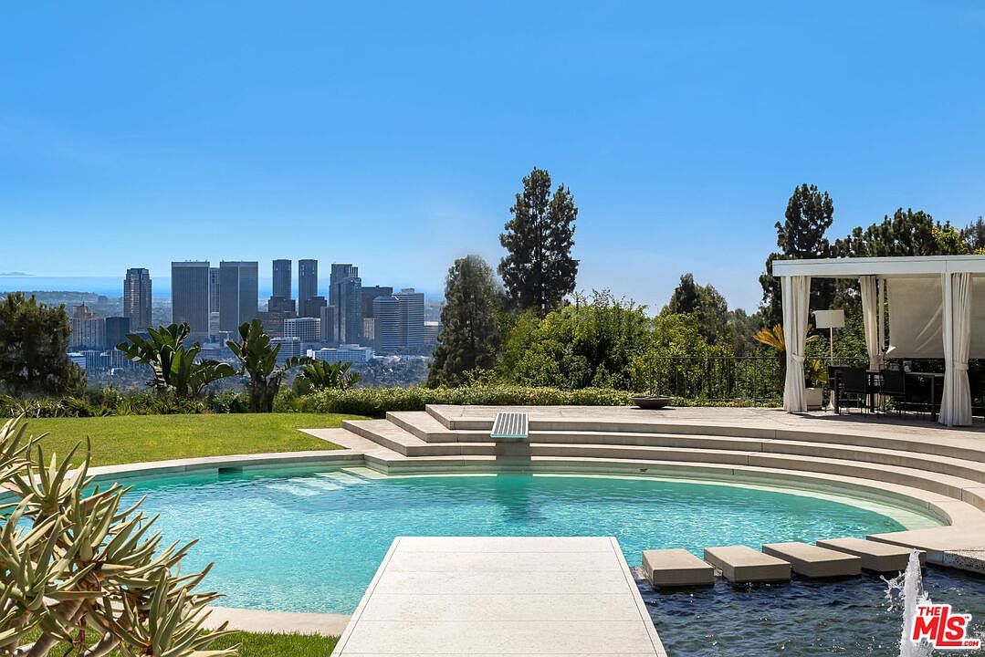 This image showcases a luxurious outdoor pool area with a stunning city view. The pool features a unique design with stepping stones and a modern diving board, surrounded by lush greenery and a well-manicured lawn. A covered patio area with elegant furniture adds to the appeal, creating an ideal space for relaxation and entertainment.