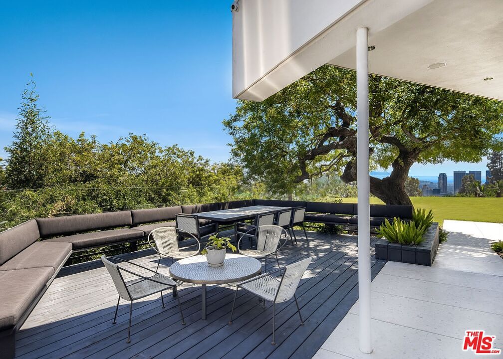 This image showcases a stylish outdoor patio or deck area, featuring modern furniture including a round table with chairs and a long bench with cushions. The deck is constructed with dark-stained wood, and the space is partially covered by an architectural overhang supported by a white pillar. Lush greenery and a view of the city skyline add to the appeal of this outdoor living space.
