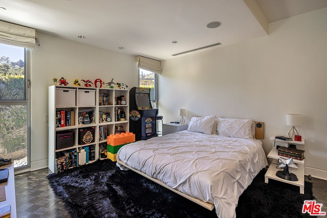 This is a cozy bedroom featuring a bed with white bedding, flanked by a white nightstand on one side and a bookshelf on the other. An arcade game stands next to the bookshelf, adding a playful touch. The room is illuminated by natural light from a window and a black shag rug adds texture to the floor.