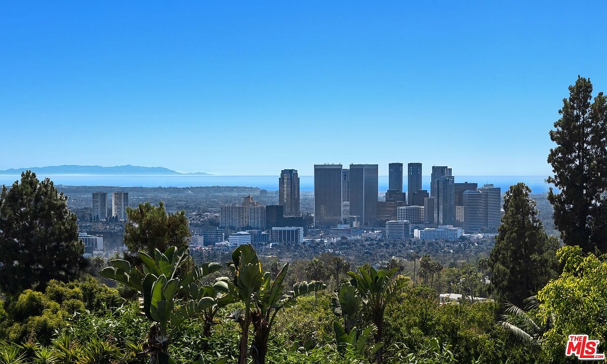 This aerial view showcases a cityscape with numerous high-rise buildings against a clear blue sky and distant ocean horizon. Lush greenery in the foreground provides a natural contrast to the urban landscape, creating a sense of tranquility and seclusion. The perspective highlights the property's proximity to both city amenities and natural beauty.