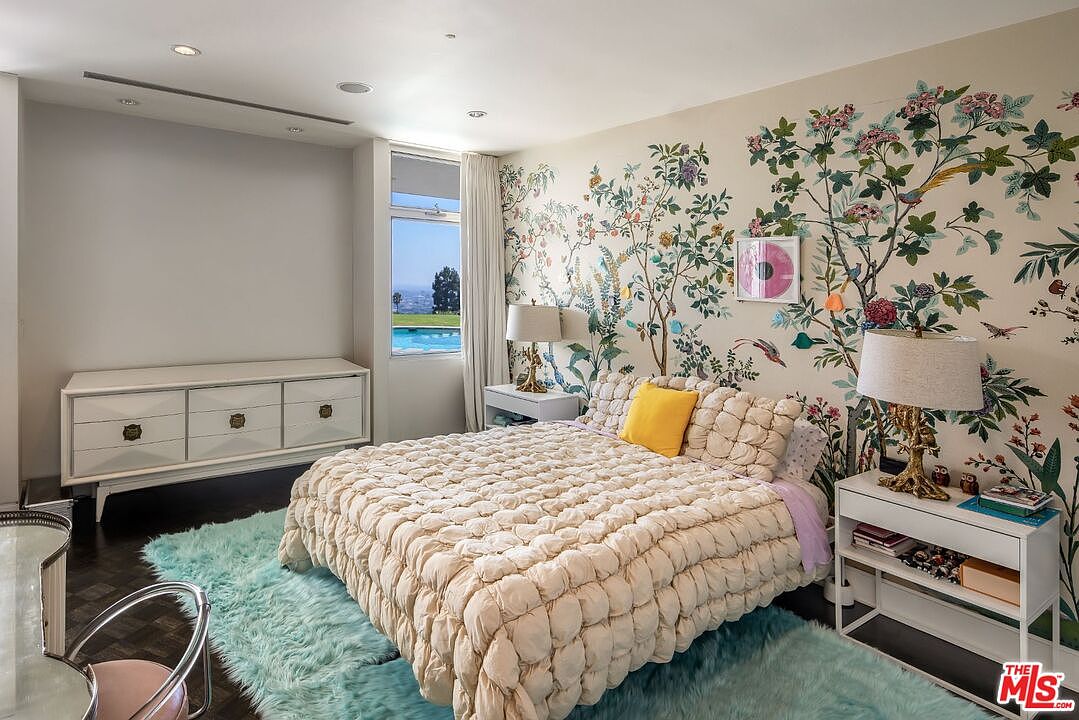 This is a primary bedroom featuring a large bed with a puffy comforter and a yellow accent pillow. The room has a floral wallpaper accent wall and a white dresser. A window offers a view of the pool and landscape, and a light blue rug covers the floor.