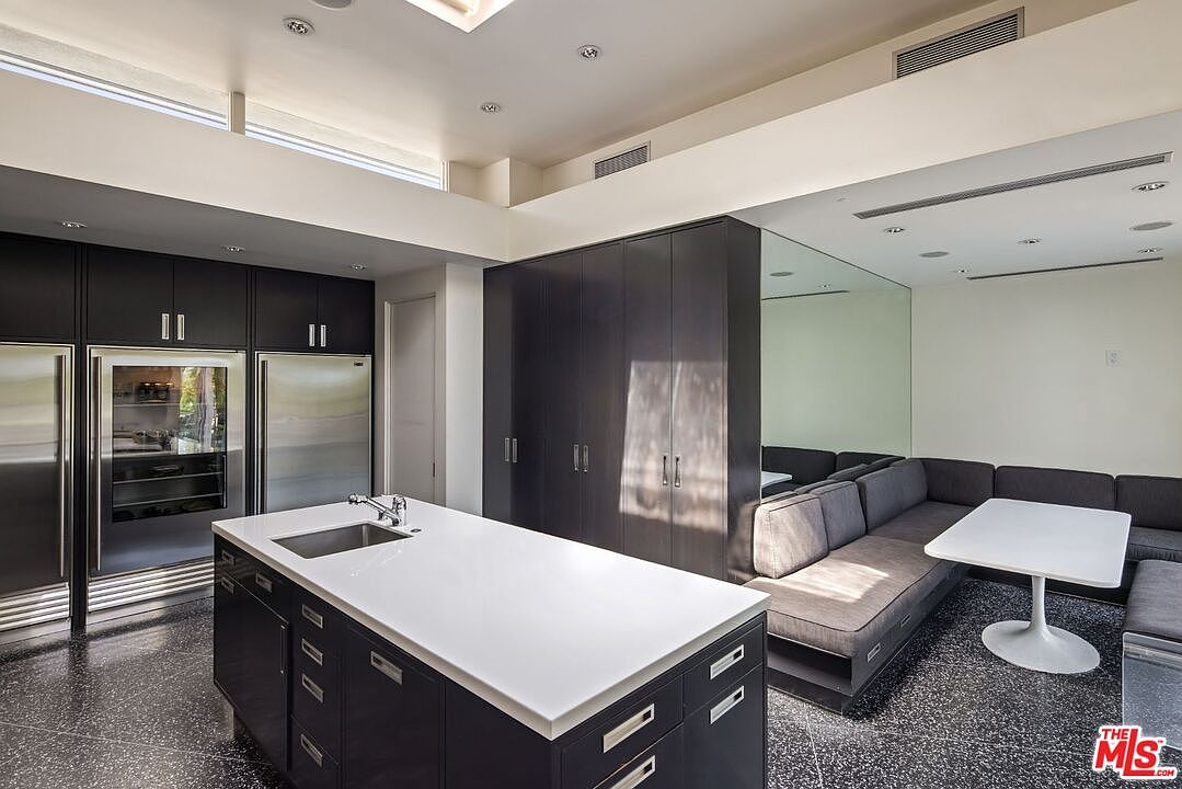 This is a modern kitchen with dark cabinetry, stainless steel appliances, and a white countertop island. The kitchen seamlessly transitions into a living area with a gray sectional sofa and a white tulip table, creating an open and inviting space. The flooring is a speckled dark material, and the overall design is sleek and contemporary.