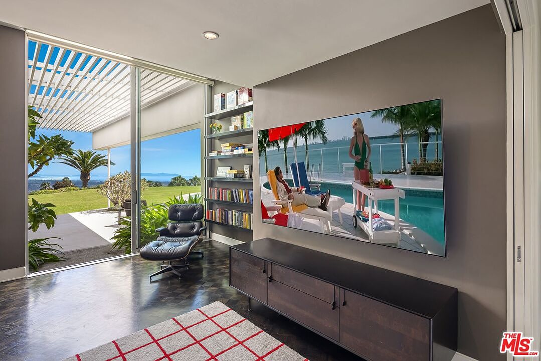 This living room features a large sliding glass door with a view of the ocean, a modern entertainment center with a large television, and a comfortable leather chair. A bookshelf is built into the wall, and a patterned rug adds warmth to the dark wood floors. The room has a sleek, contemporary design with a focus on natural light and the stunning outdoor scenery.