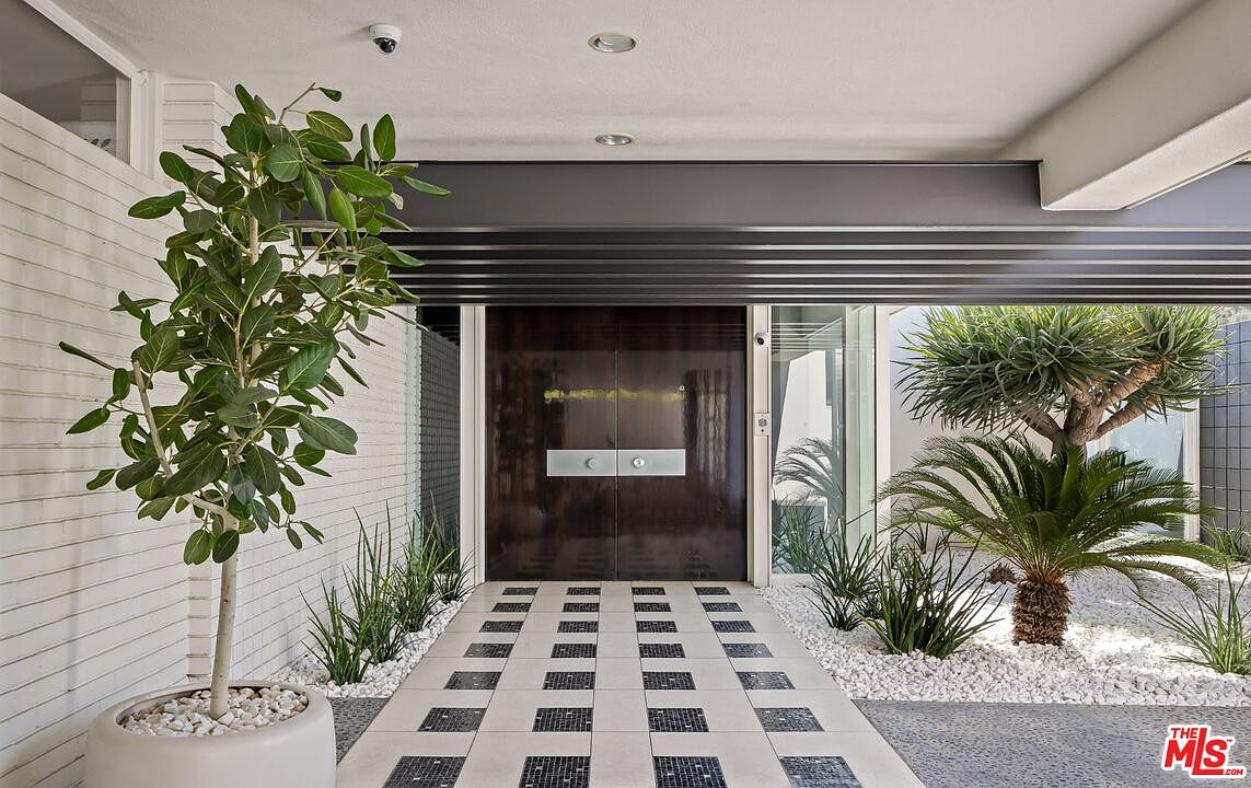 This is a modern entryway featuring a pair of dark wood double doors with silver hardware, flanked by lush greenery and white gravel. The walkway is paved with a pattern of light and dark tiles, leading to the entrance. The overall design is sleek and sophisticated, creating a welcoming and stylish first impression.