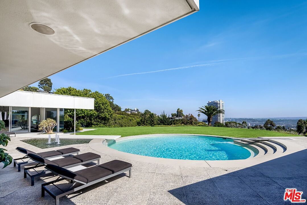 This image showcases a luxurious outdoor pool area with a modern architectural style. The pool is surrounded by a concrete patio and lush green lawn, offering a serene view of the cityscape in the distance. Lounge chairs are neatly arranged, inviting relaxation and enjoyment of the outdoor space.