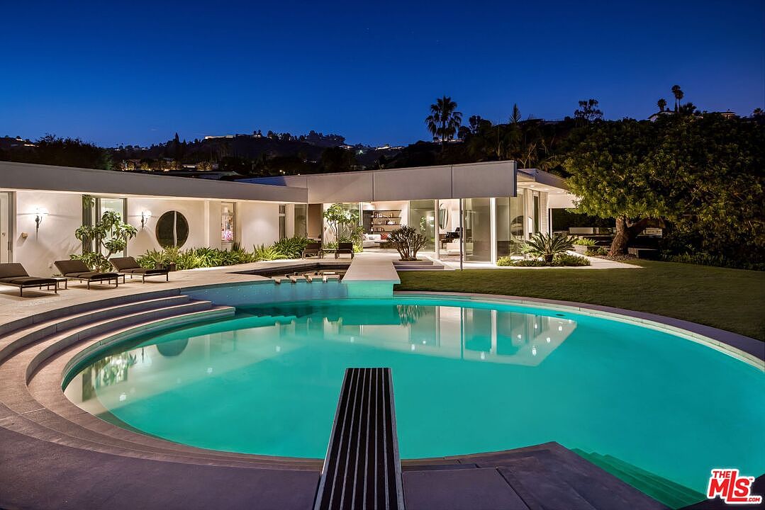 This image showcases a luxurious backyard featuring a large, circular swimming pool with a diving board. The pool is surrounded by a modern, white-walled house with expansive glass windows and doors, seamlessly blending indoor and outdoor living. Lush greenery and manicured lawns enhance the property's appeal, creating a serene and upscale atmosphere.