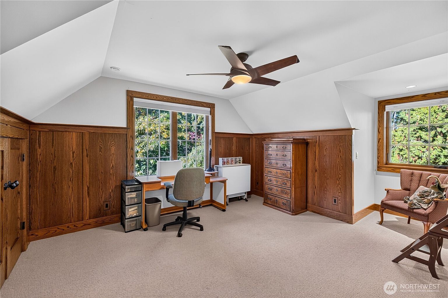 This home office features warm wood wainscoting that wraps around the lower walls, creating a classic and cozy atmosphere. The room is furnished with a desk, an office chair, a tall wooden dresser, and a comfortable armchair, all set against a neutral carpeted floor. Large windows allow natural light to fill the space, which is topped by a vaulted ceiling with a central wooden ceiling fan.