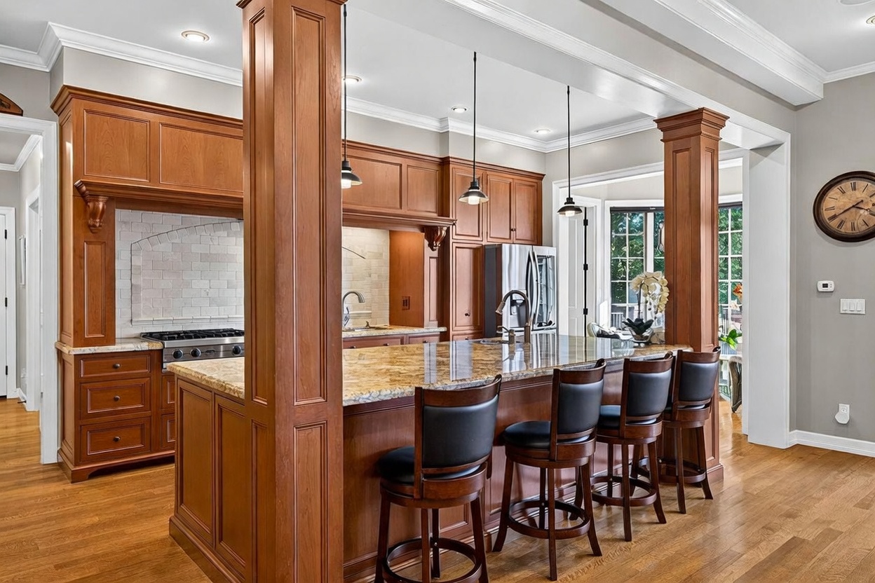 This spacious kitchen features warm-toned wood cabinetry, a large central island with a stone countertop, and four bar stools. The space is defined by two prominent wooden columns and is illuminated by three pendant lights hanging over the island. A light-colored tiled backsplash sits behind the range, and a large wall clock is mounted on the right wall, creating a traditional and inviting atmosphere.