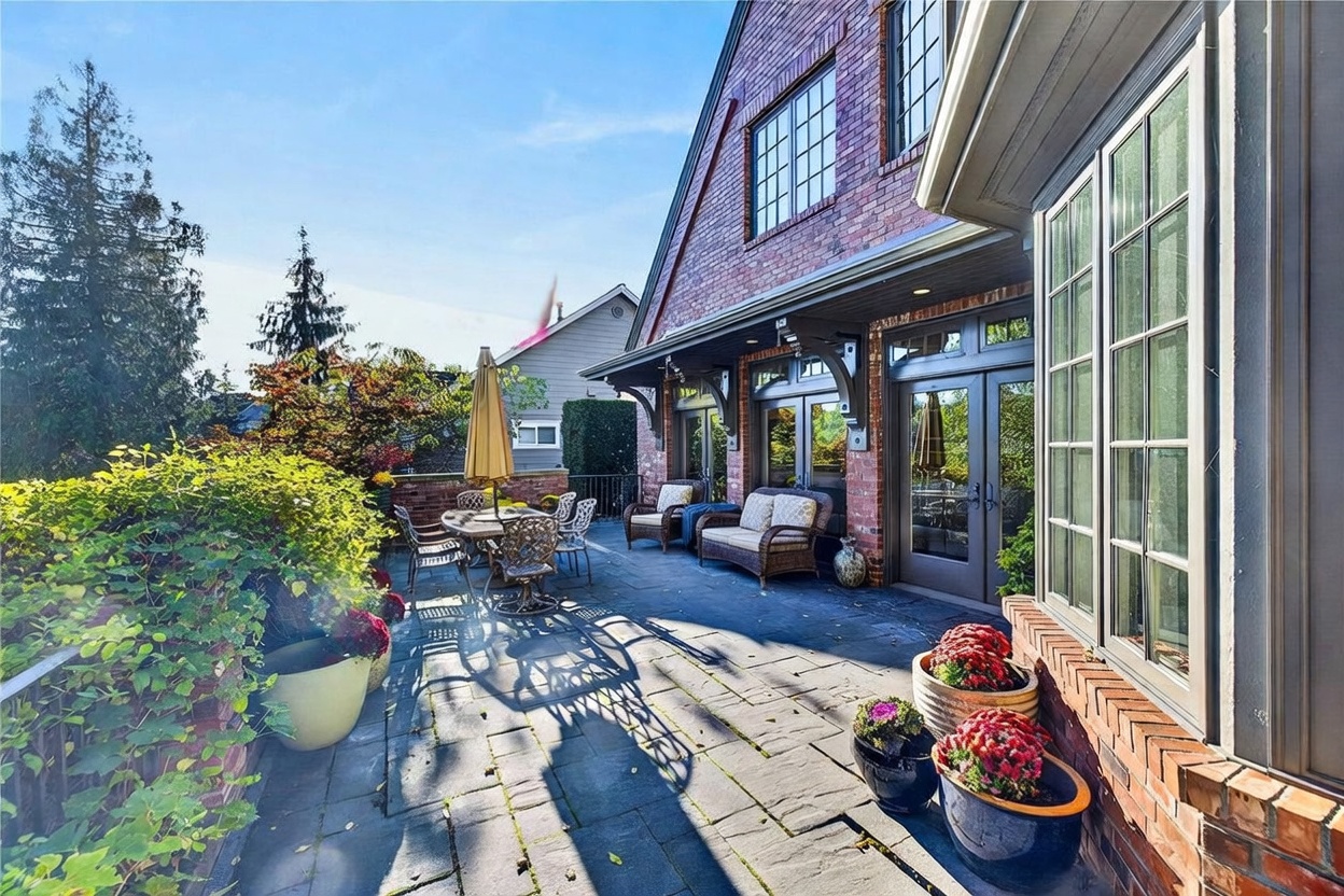 This inviting brick-clad patio offers a spacious outdoor living area, complete with a dining set and comfortable wicker seating. The space is accented by lush potted plants and vibrant flowers, creating a warm and welcoming atmosphere against the backdrop of the home's classic architecture. Large glass doors provide a seamless transition between the indoor and outdoor living spaces, perfect for entertaining.