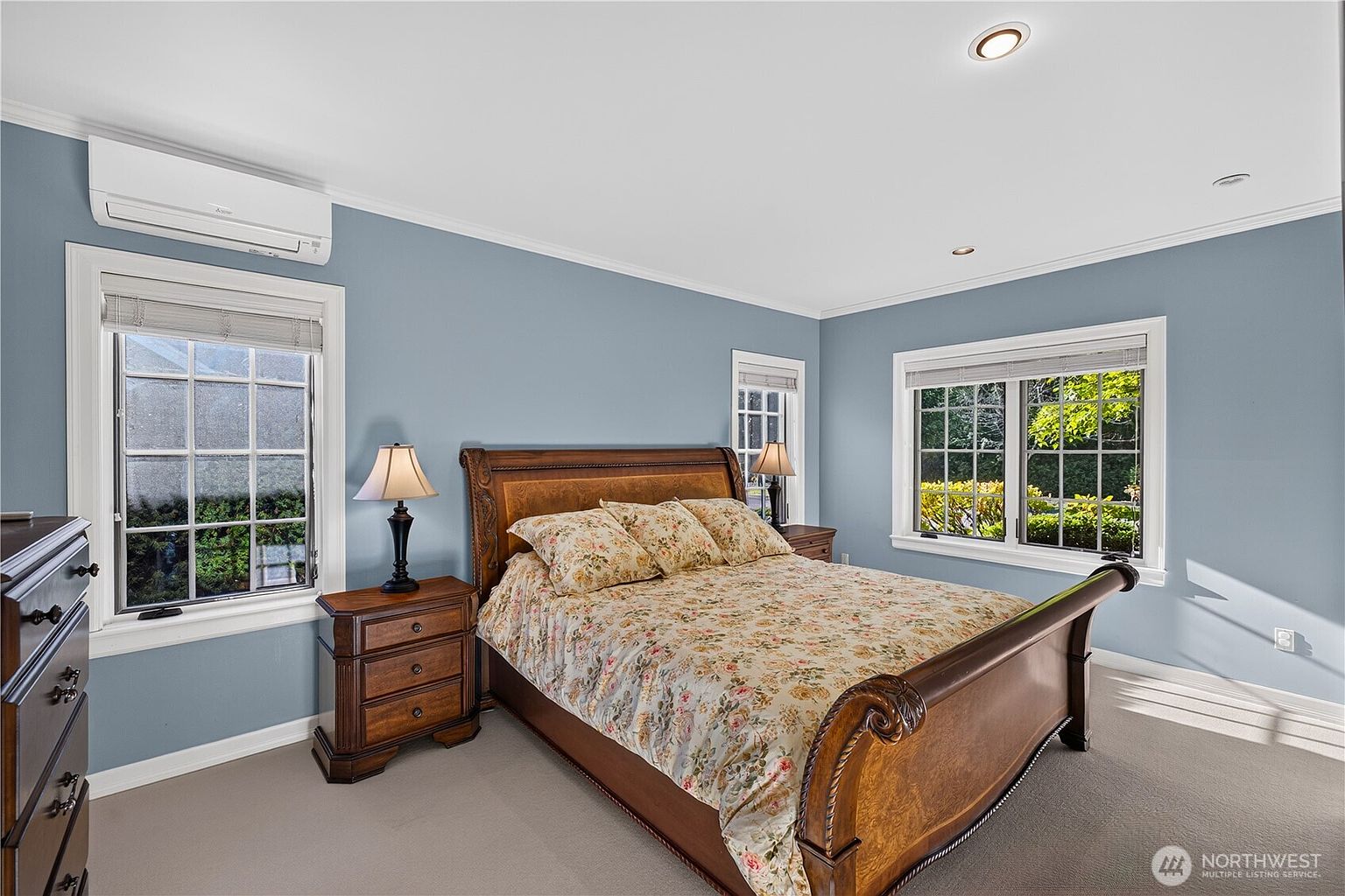 This primary bedroom features light blue walls and neutral carpeting, creating a calm and inviting atmosphere. A substantial wooden sleigh bed with a floral-patterned comforter serves as the focal point, flanked by matching wooden nightstands and lamps. The room is well-lit by natural light streaming through two windows and includes a wall-mounted mini-split air conditioning unit.