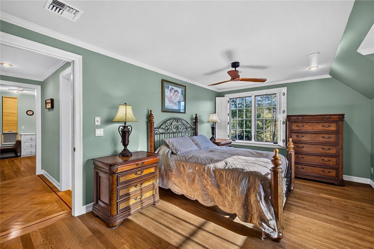 This bedroom features sage green walls, warm-toned wood flooring, and a classic metal-framed bed as the central focal point. The room is furnished with matching dark wood dressers and bedside tables, complemented by a ceiling fan and a window with white shutters that allows natural light to enter. A doorway on the left provides a glimpse into an adjacent bathroom, creating a cohesive and traditional living space.