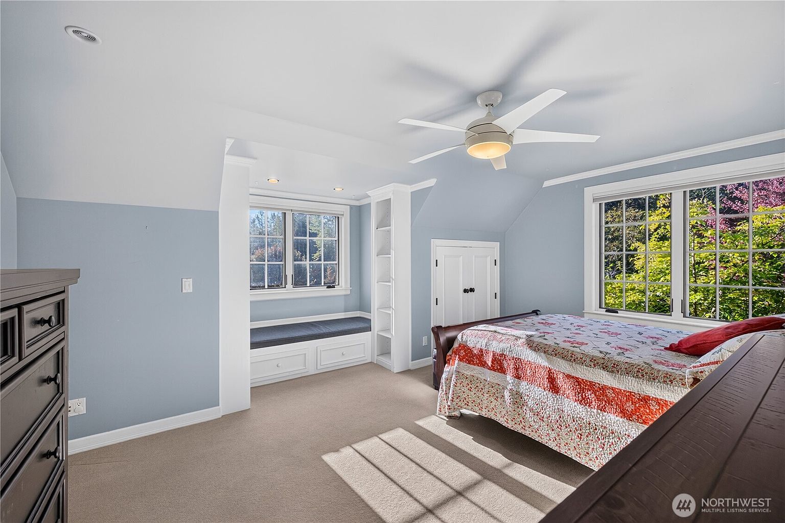 This bedroom features light blue walls, neutral carpeting, and a cozy built-in window seat flanked by white shelving. A bed with a patterned quilt sits under a ceiling fan, while a window provides a view of the surrounding trees. The room has a bright, airy feel with a sloped ceiling that adds architectural character.