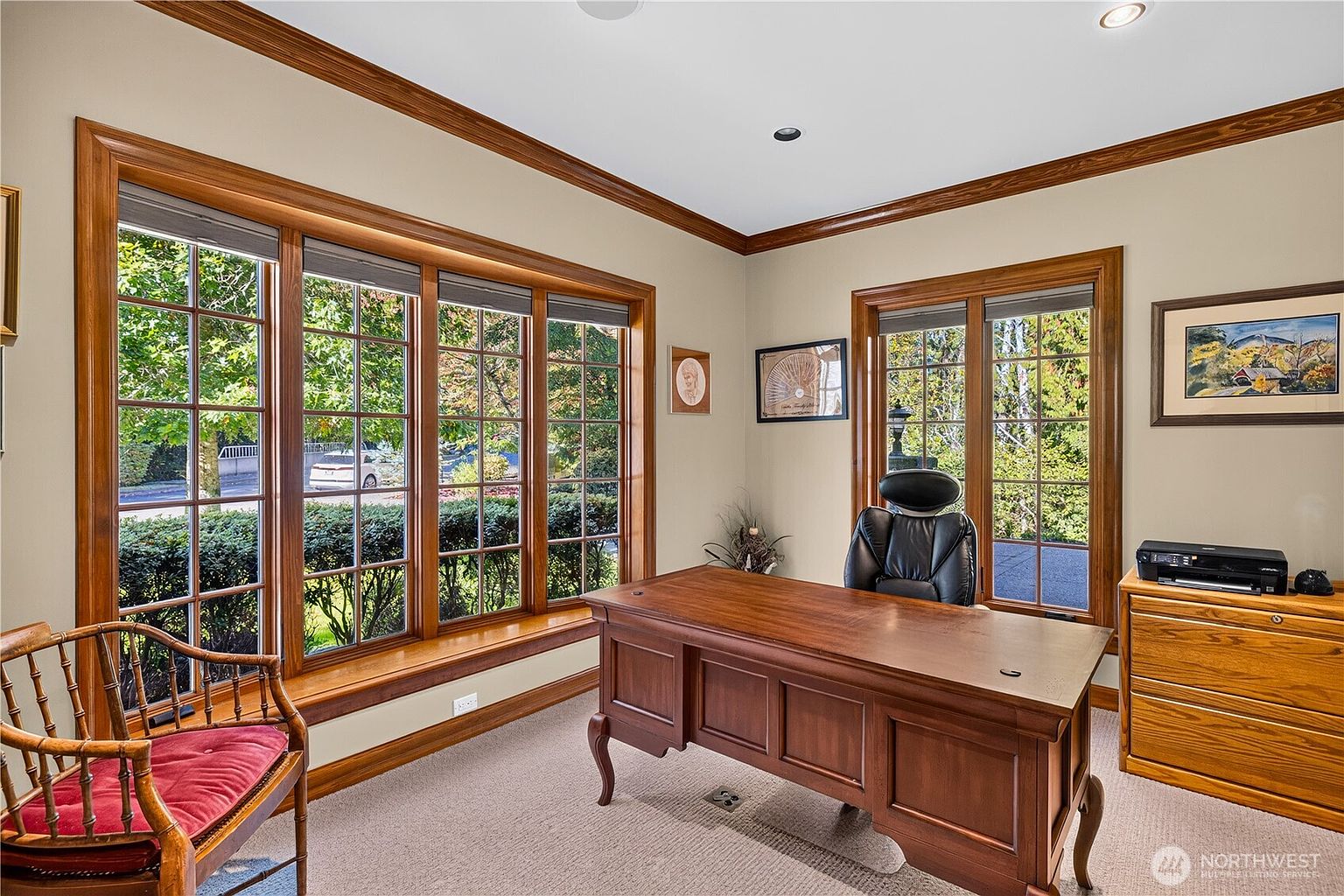This home office features a classic, traditional aesthetic with rich wood trim, crown molding, and neutral wall tones. A large wooden desk is positioned centrally, complemented by a black leather office chair and a wooden spindle chair with a red cushion. The room is brightened by multiple large windows that provide a view of the exterior landscaping.