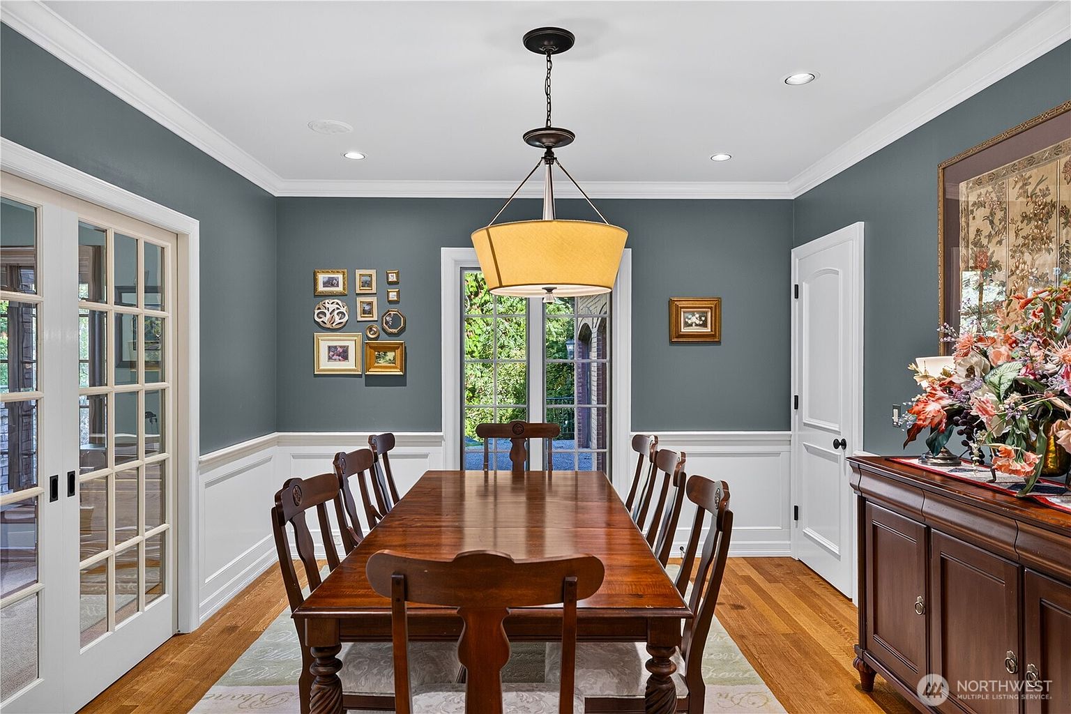 This formal dining room features dark blue walls contrasted by crisp white wainscoting and trim. A large wooden dining table with matching chairs sits centered under a drum-style pendant light, while a sideboard with a floral arrangement provides additional storage. The room is accented by a gallery wall of framed art and glass-paned doors that allow natural light to enter the space.