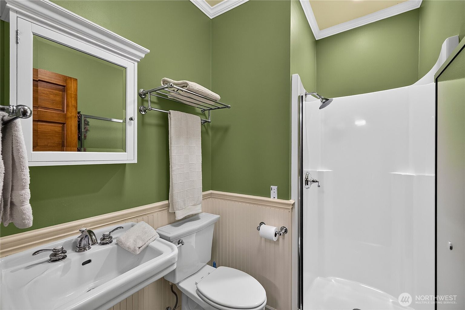 This compact bathroom features vibrant green walls contrasted by white wainscoting and a white pedestal sink. A chrome towel rack is mounted above the toilet, and a white shower stall with a chrome fixture is positioned to the right. The room is illuminated by a mirror-fronted medicine cabinet and overhead lighting, creating a clean and functional space.
