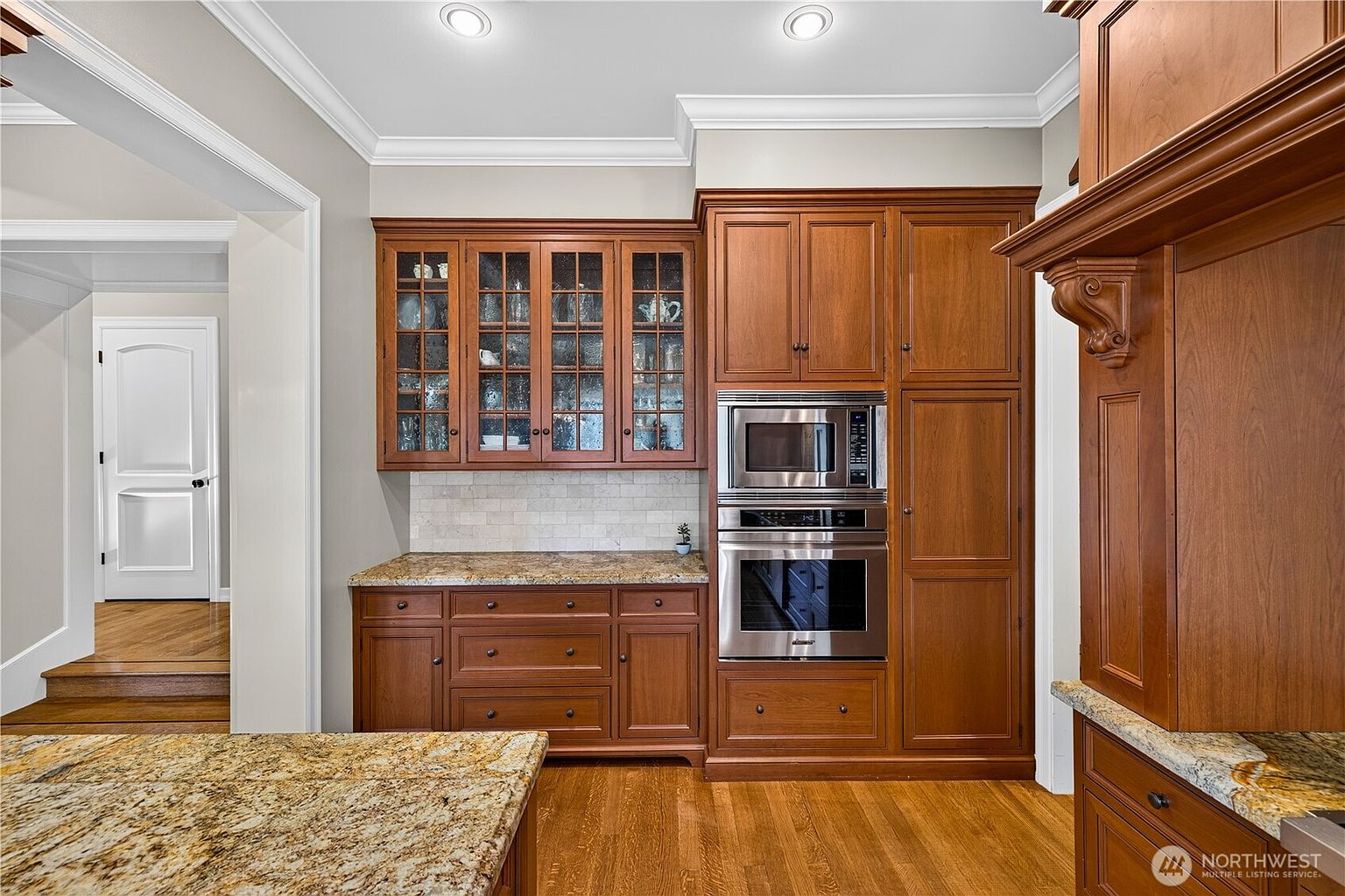 This kitchen features rich, warm-toned cabinetry with glass-front upper cabinets and a built-in stainless steel microwave and oven unit. The space is accented by a light-colored stone countertop and a neutral tiled backsplash, creating a classic and elegant aesthetic. A doorway in the background leads to a hallway with hardwood flooring, providing a sense of depth and flow to the home's layout.