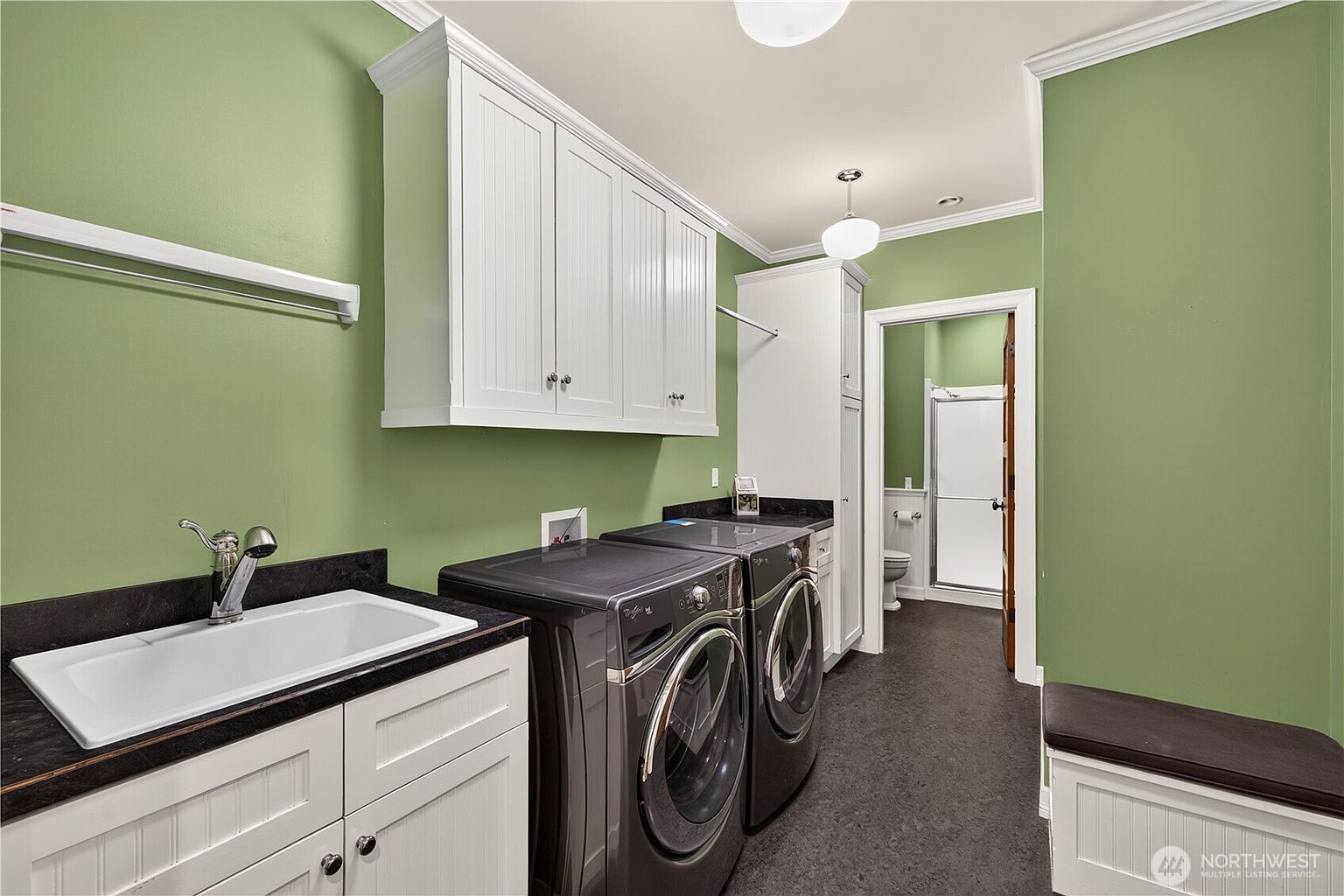 This functional laundry room features sage green walls, white cabinetry, and dark countertops. The space is equipped with a utility sink, a side-by-side washer and dryer set, and a built-in bench with storage. A doorway leads to an adjacent bathroom, and the room is illuminated by two overhead pendant lights.