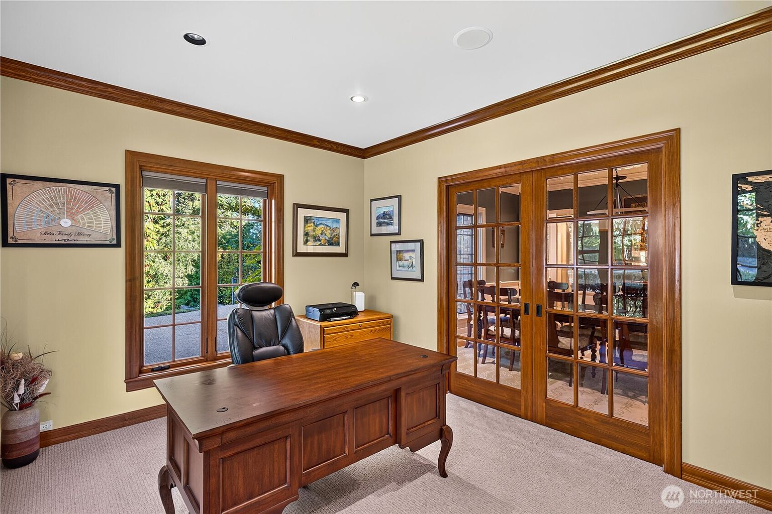 This home office features a traditional aesthetic with warm-toned wood trim, a large wooden desk, and a black leather executive chair. The room is illuminated by recessed lighting and natural light streaming through a large window, while French doors with glass panes provide a view into the adjacent dining area. The space is decorated with framed artwork and a family history chart, creating a professional yet inviting atmosphere.