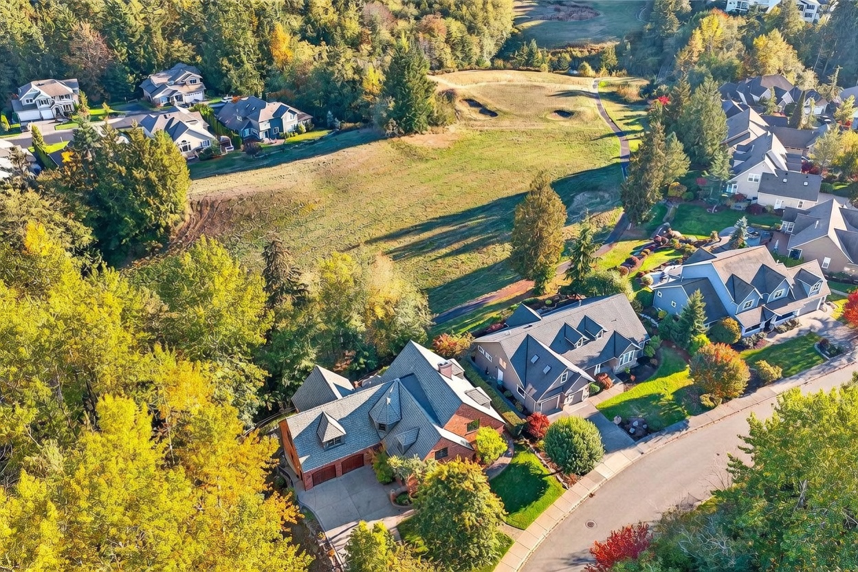 This high-angle aerial view captures a suburban neighborhood nestled against a large, open grassy field and dense woodland. The perspective highlights the layout of several multi-story homes with complex rooflines, brick and siding exteriors, and attached garages. The scene is bathed in warm, golden-hour sunlight, emphasizing the lush green lawns and the surrounding mature trees during the autumn season.