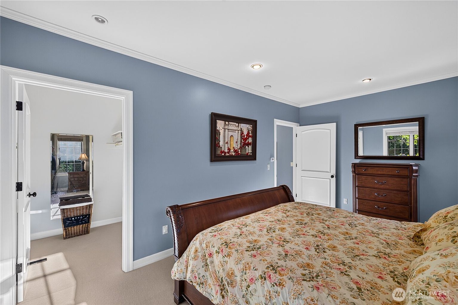 This primary bedroom features soft blue walls and a large, dark wood sleigh bed adorned with a floral-patterned comforter. A matching dark wood dresser with a mirror sits against the wall, while an open doorway leads into a walk-in closet with a full-length mirror. The room is illuminated by recessed lighting, creating a calm and traditional atmosphere.