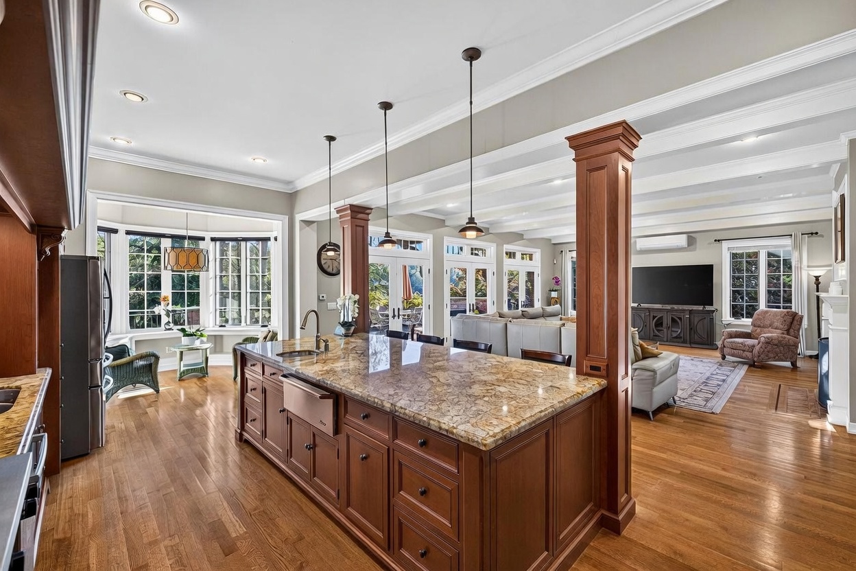 This open-concept kitchen features a large central island with a stone countertop and a farmhouse-style sink, serving as the focal point of the room. The space is anchored by rich wood cabinetry and warm-toned hardwood flooring, with pendant lighting hanging above the island. The kitchen flows seamlessly into a bright living area, creating an expansive and inviting atmosphere suitable for entertaining.