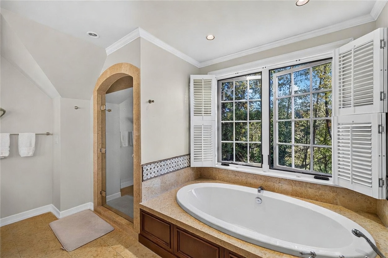 This primary bathroom features a deep soaking tub set into a stone-topped vanity with dark wood cabinetry. A glass-enclosed shower stall is framed by an arched stone entryway, and large windows with white shutters provide natural light and a view of the surrounding trees. The space is finished with neutral wall tones and patterned tile accents, creating a serene and classic atmosphere.