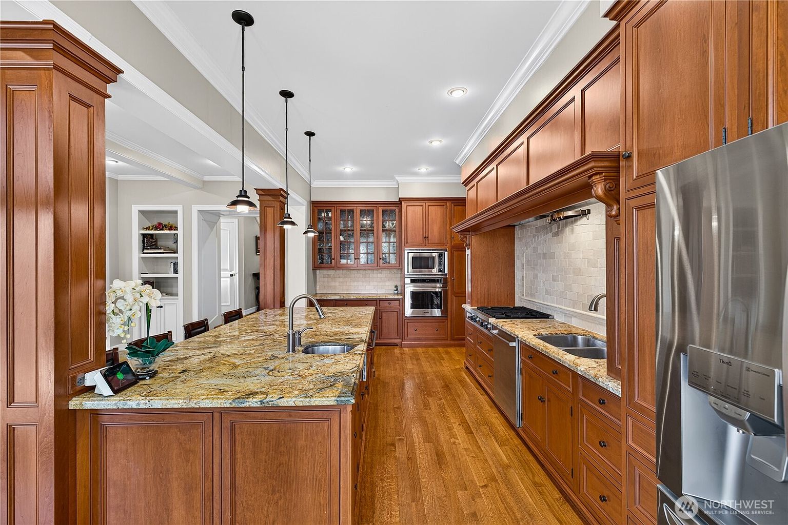 This spacious kitchen features warm-toned wood cabinetry, a large central island with a stone countertop, and stainless steel appliances. The layout includes a dedicated cooking area with a tiled backsplash and a secondary sink, all illuminated by recessed lighting and three pendant lights hanging over the island. The perspective is a wide-angle shot from the kitchen entrance, showcasing the open flow and the rich, traditional aesthetic of the cabinetry and hardwood flooring.