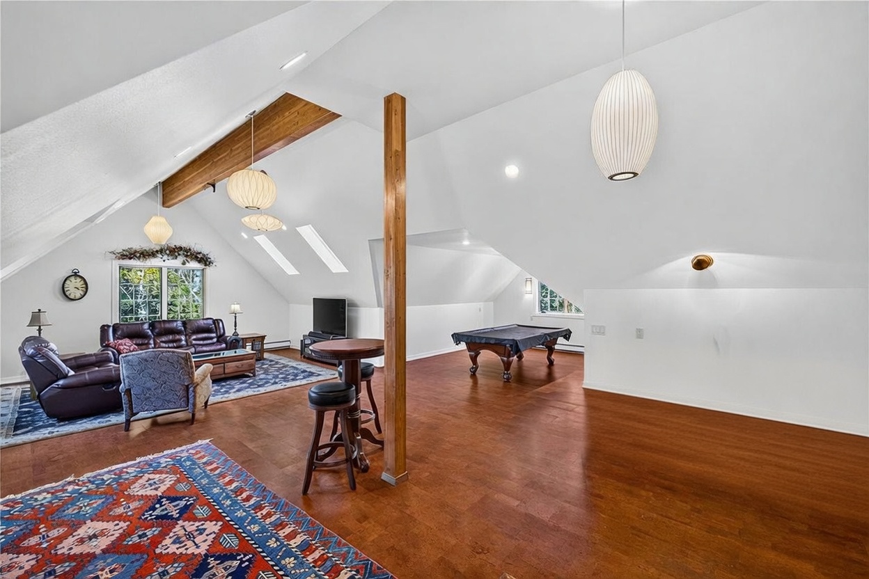 This expansive, open-concept attic space features vaulted ceilings with exposed wooden beams and multiple skylights that flood the room with natural light. The area is multi-functional, incorporating a cozy living room with leather seating, a central bar area with high-top stools, and a dedicated space for a pool table. The warm wood flooring and neutral walls create a versatile and inviting recreational environment.