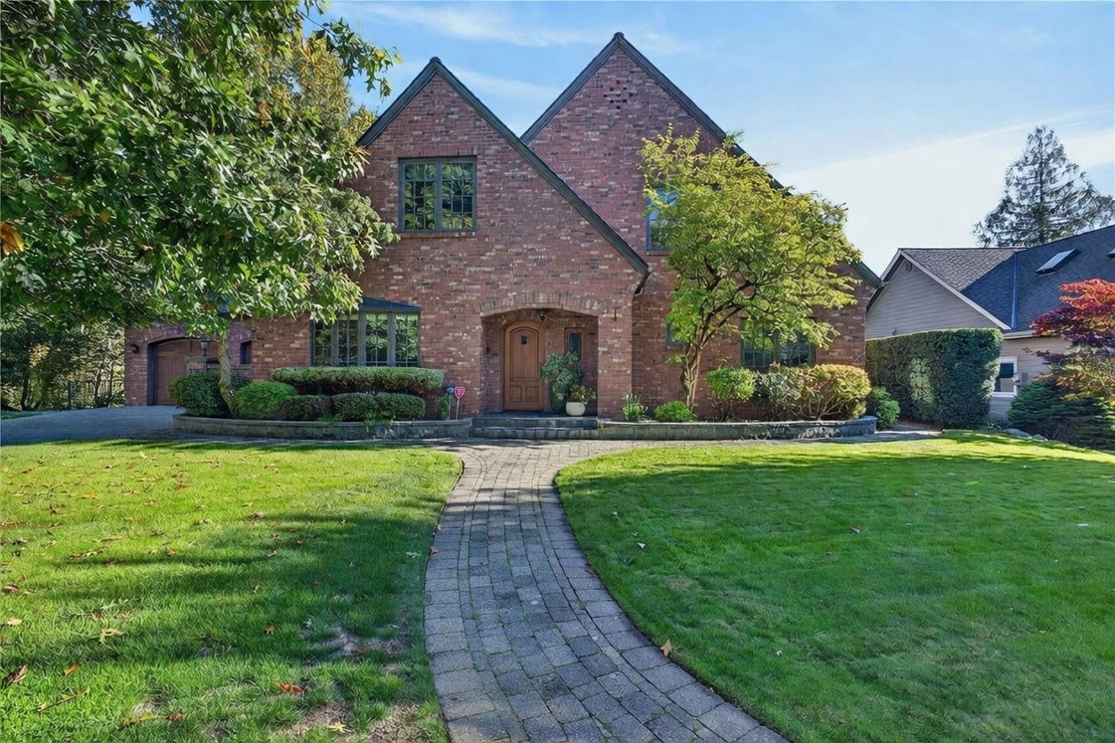 This two-story brick home features a classic architectural style with a prominent gabled roofline and a central arched entryway. A stone-paved walkway leads through a well-manicured lawn to the front door, framed by mature trees and neatly trimmed landscaping. The property presents a welcoming and established curb appeal, captured from a straight-on perspective.