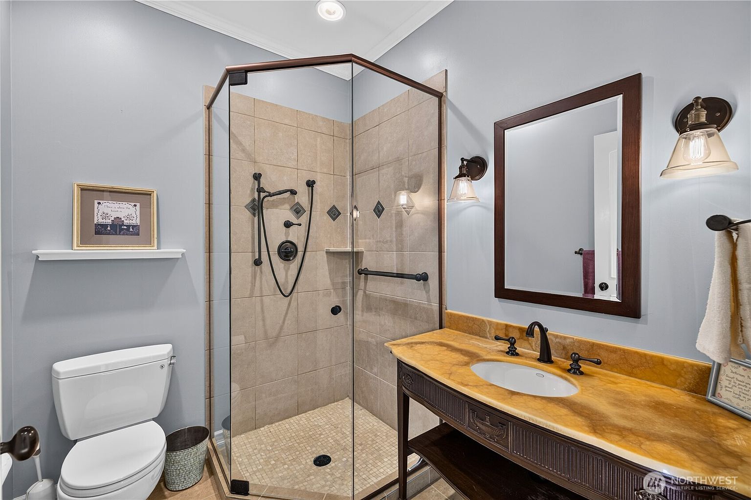 This bathroom features a corner glass-enclosed shower with neutral-toned wall tiles and a mosaic floor. A vanity with a warm-toned stone countertop and a dark wood base sits beside a framed mirror and wall-mounted light fixtures. The room is finished with light blue walls, a white toilet, and a small decorative shelf, creating a functional and tidy space.