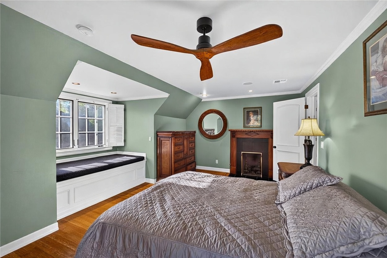This primary bedroom features sage green walls, hardwood flooring, and a cozy window seat tucked into a dormer alcove. A dark wood-framed fireplace with a decorative mantel serves as a focal point, complemented by a ceiling fan with wooden blades and a wrought-iron bed frame. The room offers a warm, traditional aesthetic with a comfortable and inviting atmosphere.