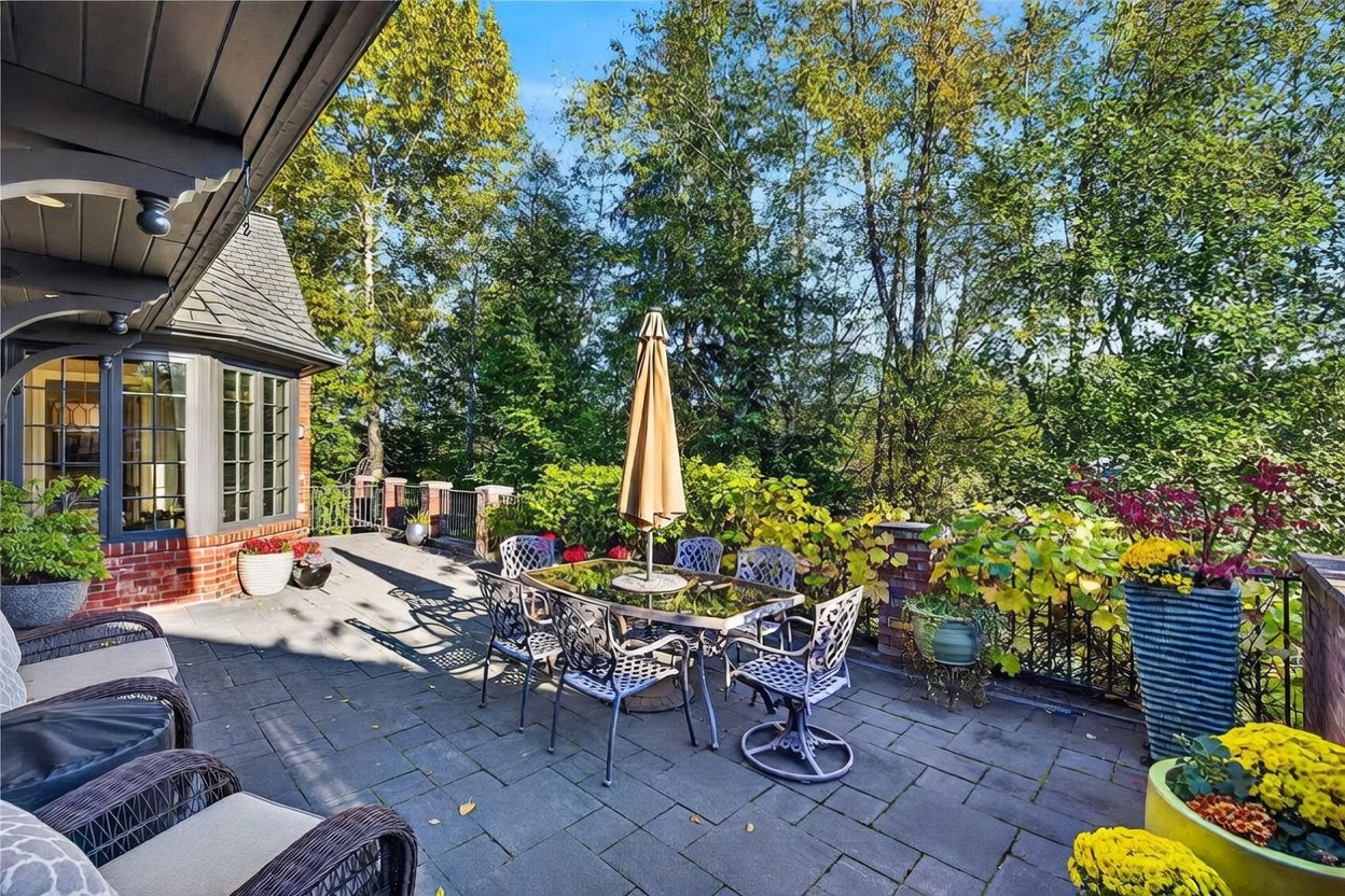 This spacious stone-paved patio offers a serene outdoor living area, complete with a dining set shaded by a tan umbrella. The space is bordered by a brick and metal railing, overlooking a lush, tree-filled backdrop that provides significant privacy. The perspective captures the transition from the home's brick exterior to the open-air terrace, creating an inviting atmosphere for relaxation or al fresco dining.