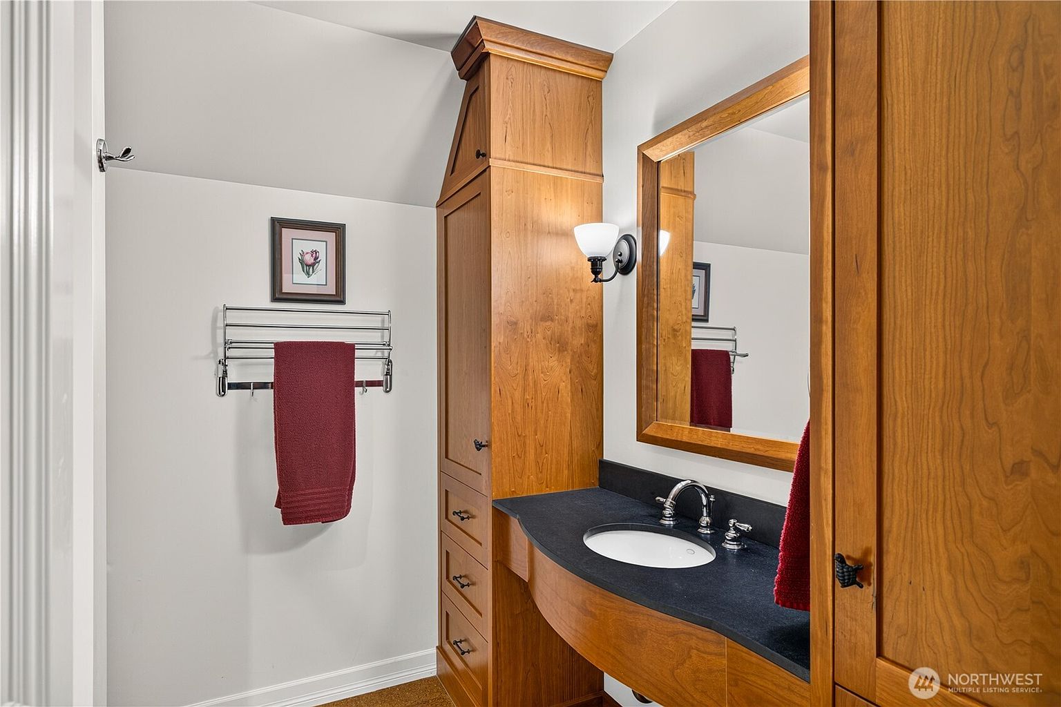 This bathroom features a custom wood vanity with a dark stone countertop and an integrated white sink. A tall, matching wood storage cabinet stands beside the vanity, complemented by a framed mirror and a wall-mounted towel rack holding a red towel. The room is finished with white walls and a sloped ceiling, creating a cozy and functional space.