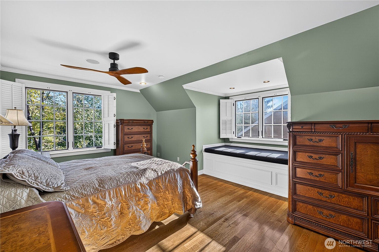 This cozy bedroom features sage green walls, warm wood flooring, and a classic wooden bed frame with a quilted bedspread. A built-in window seat with dark cushions sits beneath a dormer window, providing a charming nook, while two large wooden dressers offer ample storage. The room is illuminated by a ceiling fan with wooden blades and natural light streaming through multiple windows with white shutters.