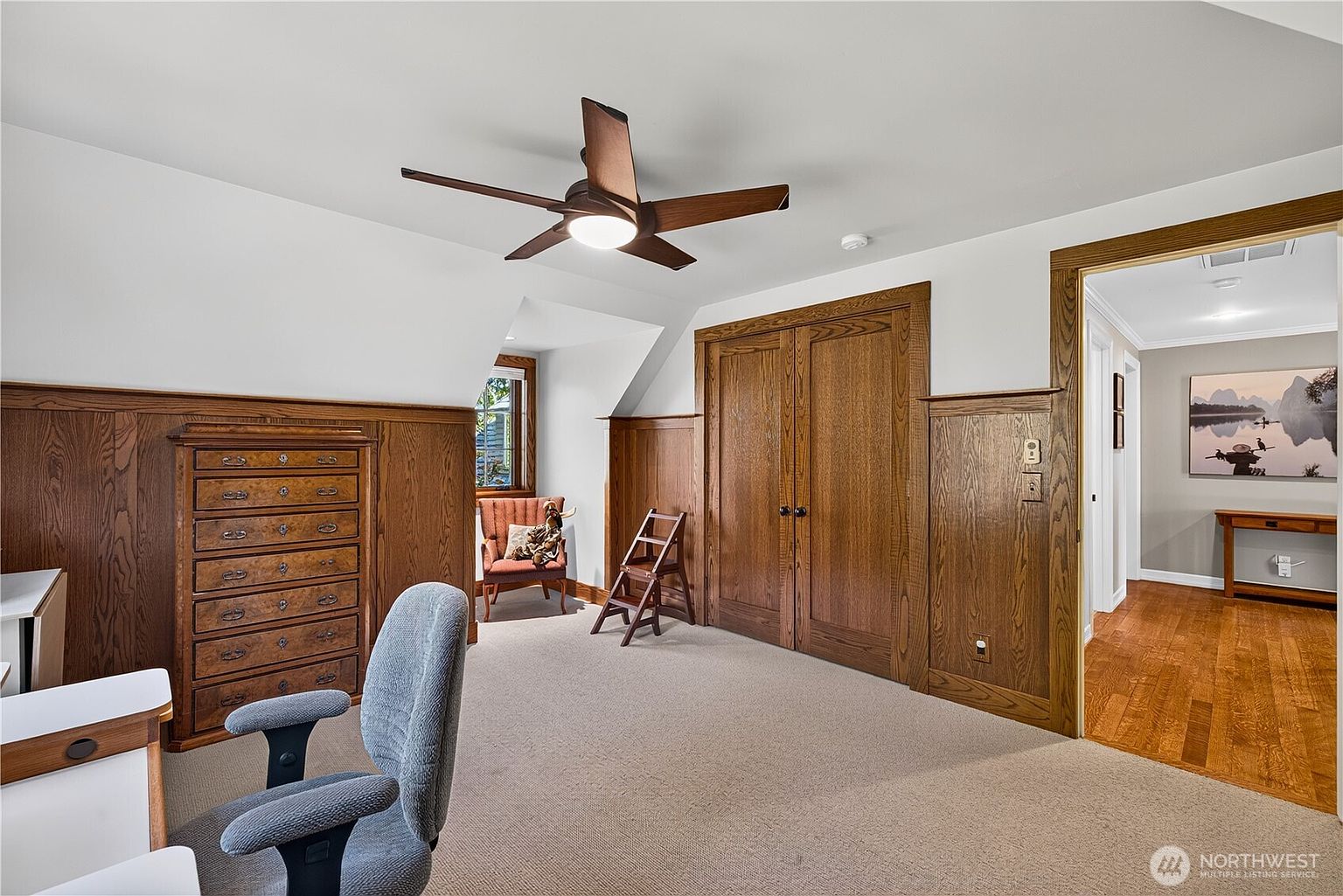 This home office features rich wood wainscoting and matching double-door closet cabinetry, creating a warm and traditional atmosphere. The room includes a desk area, a small armchair tucked into a dormer nook, and a wooden step stool, all set against a neutral carpeted floor. A ceiling fan with an integrated light fixture centers the space, while a doorway leads to an adjacent hallway with hardwood flooring.