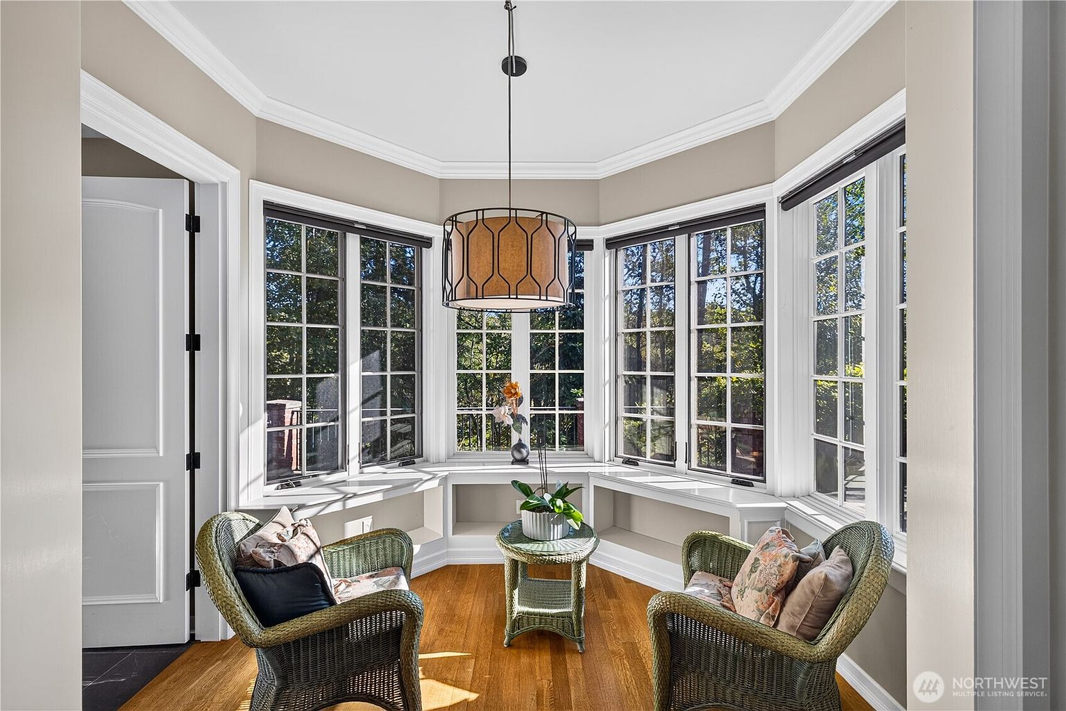 This bright sunroom features a bay window configuration with built-in bench seating and storage cubbies underneath. The space is furnished with two wicker armchairs and a matching small round table, centered beneath a decorative drum pendant light. The room has warm-toned wood flooring and neutral wall paint, creating a cozy and inviting nook with views of the surrounding trees.