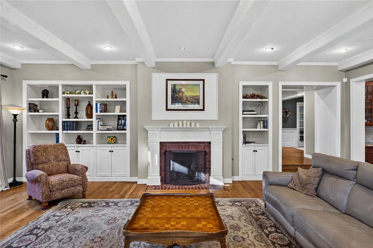 This inviting living room features a central brick fireplace with a white mantel, flanked by extensive built-in shelving and cabinetry. A patterned area rug sits beneath a wooden coffee table, while a patterned armchair and a grey sofa provide comfortable seating. The room is accented by coffered ceilings and warm wood flooring, creating a traditional and cozy atmosphere.